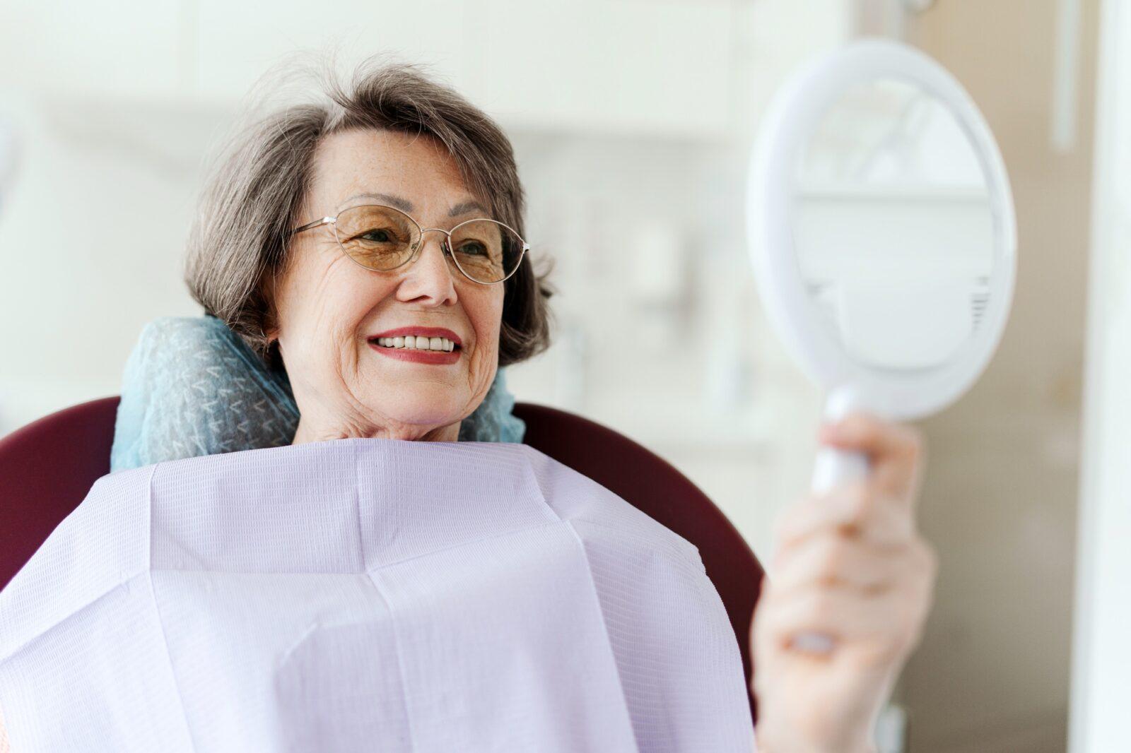 smiling-senior-female-patient-holding-mirror-looking-at-teeth-while-sitting-in-dental-chair